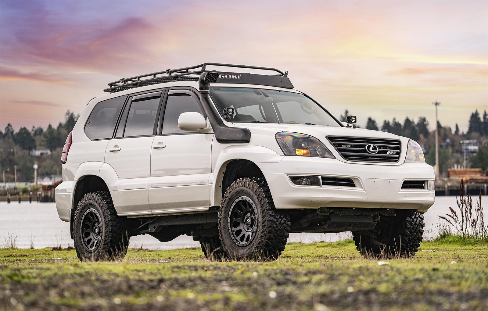 A white suv with a roof rack is parked in a grassy field next to a body of water.